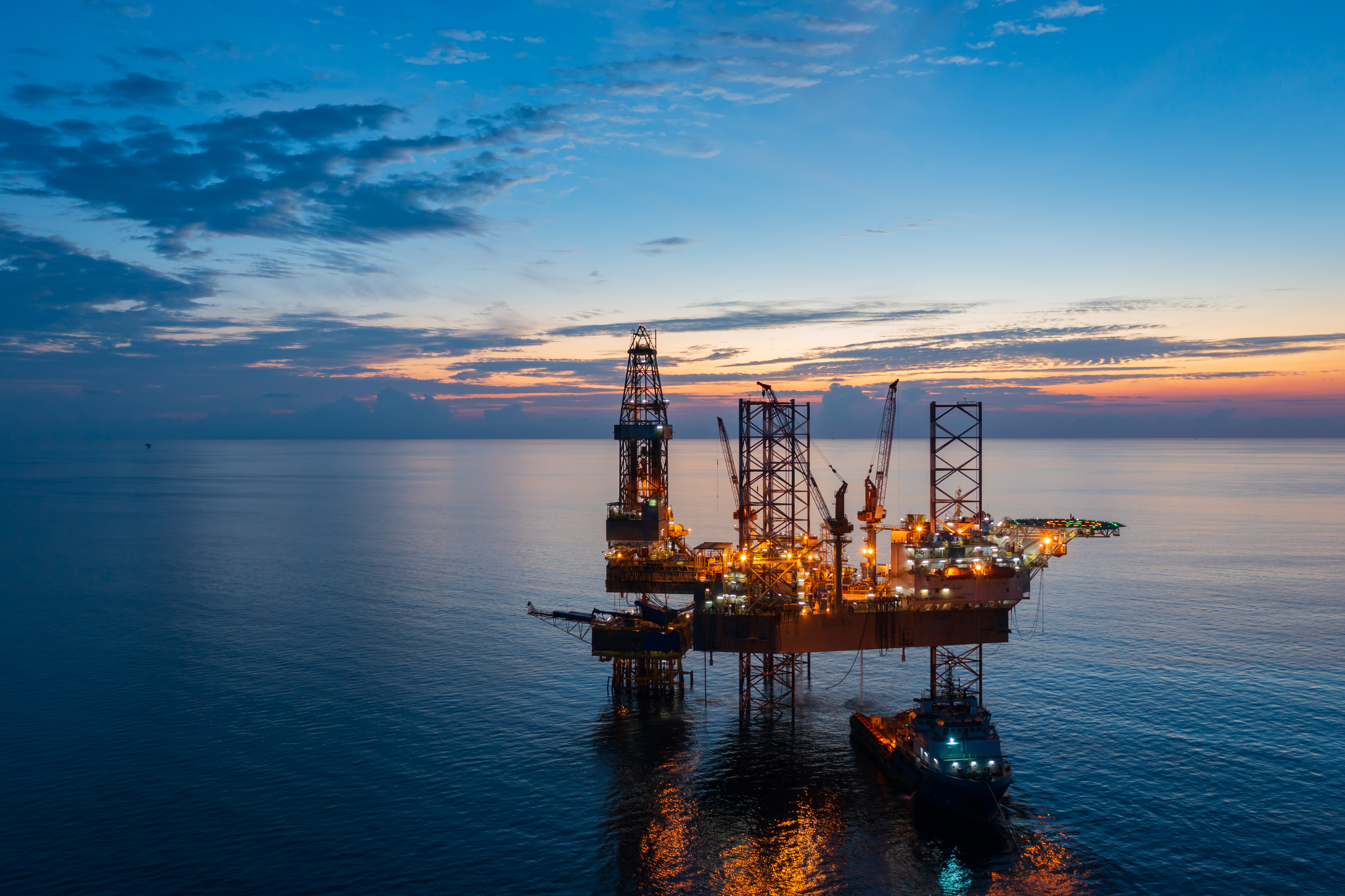 Offshore oil rig platform at sea during sunset with illuminated structures and a support vessel nearby.