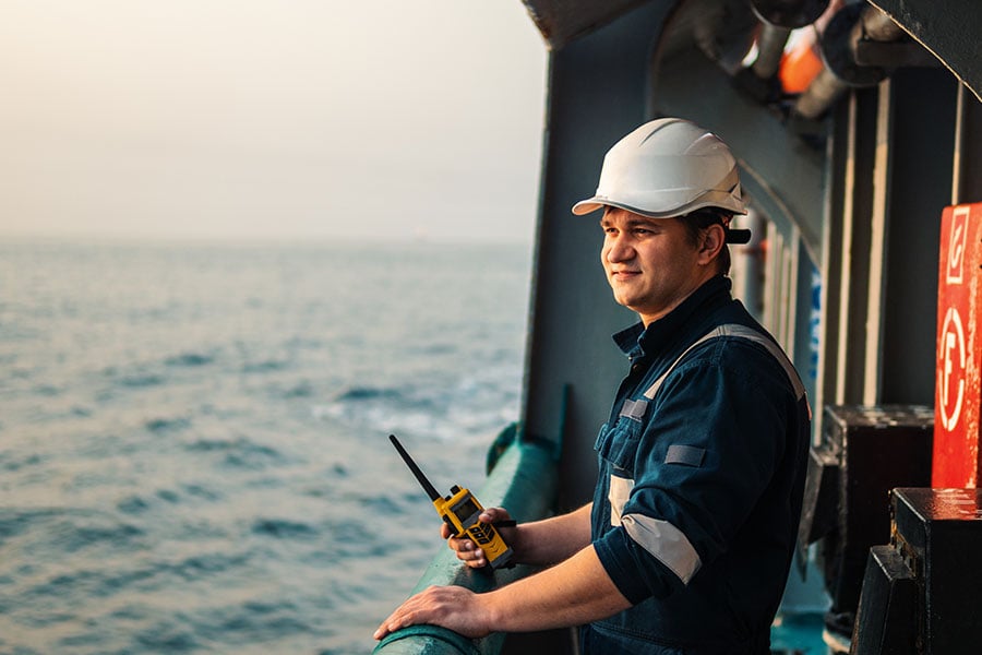 Portrait of a man in a white hard hat holding a walkie-talkie on a ship deck overlooking the ocean.