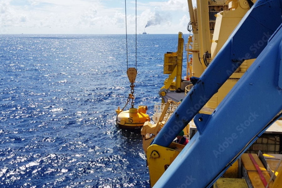 Yellow offshore rescue capsule being lowered from a ship into the ocean, with blue sea and a distant vessel on the horizon.