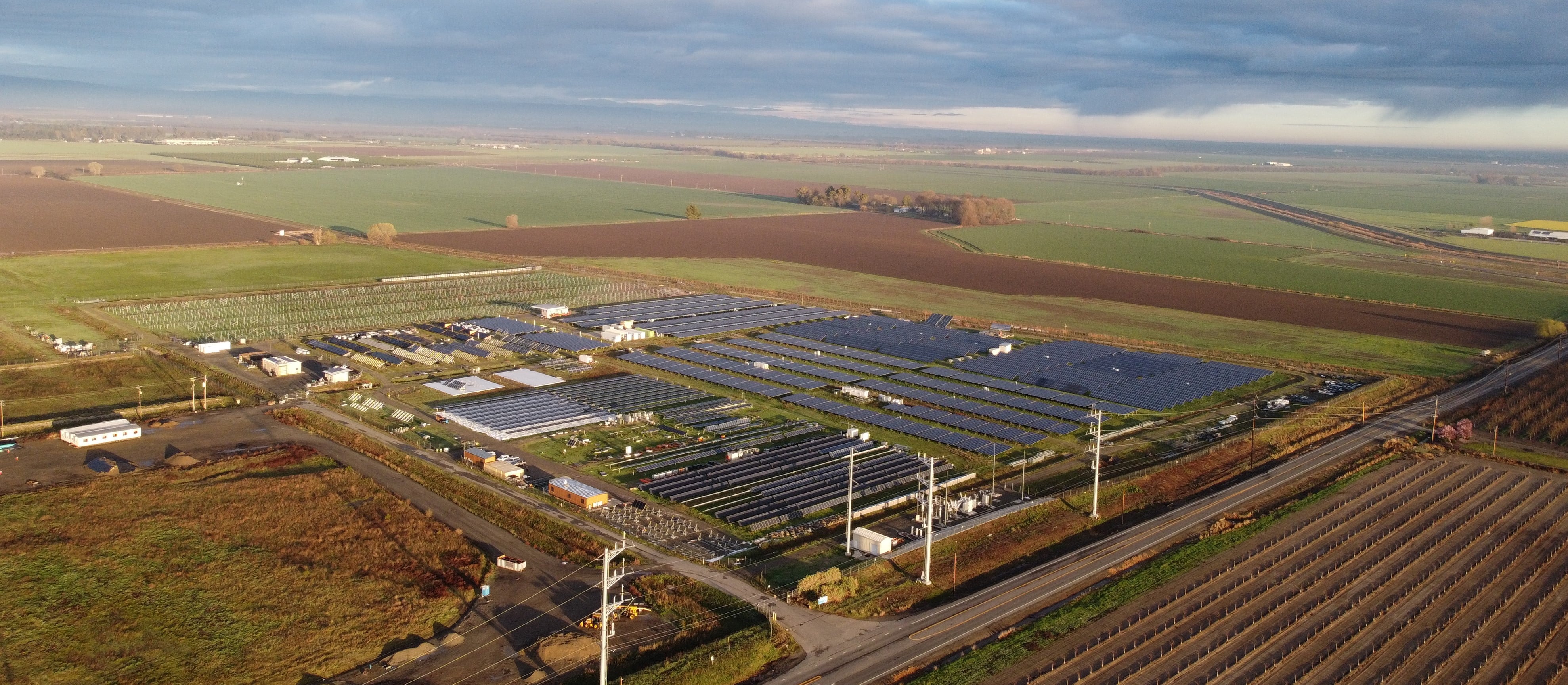 Aerial view of a solar farm with numerous solar panels spread across a vast agricultural landscape. Rolling farmland and distant roads surround the solar facility under a partly cloudy sky