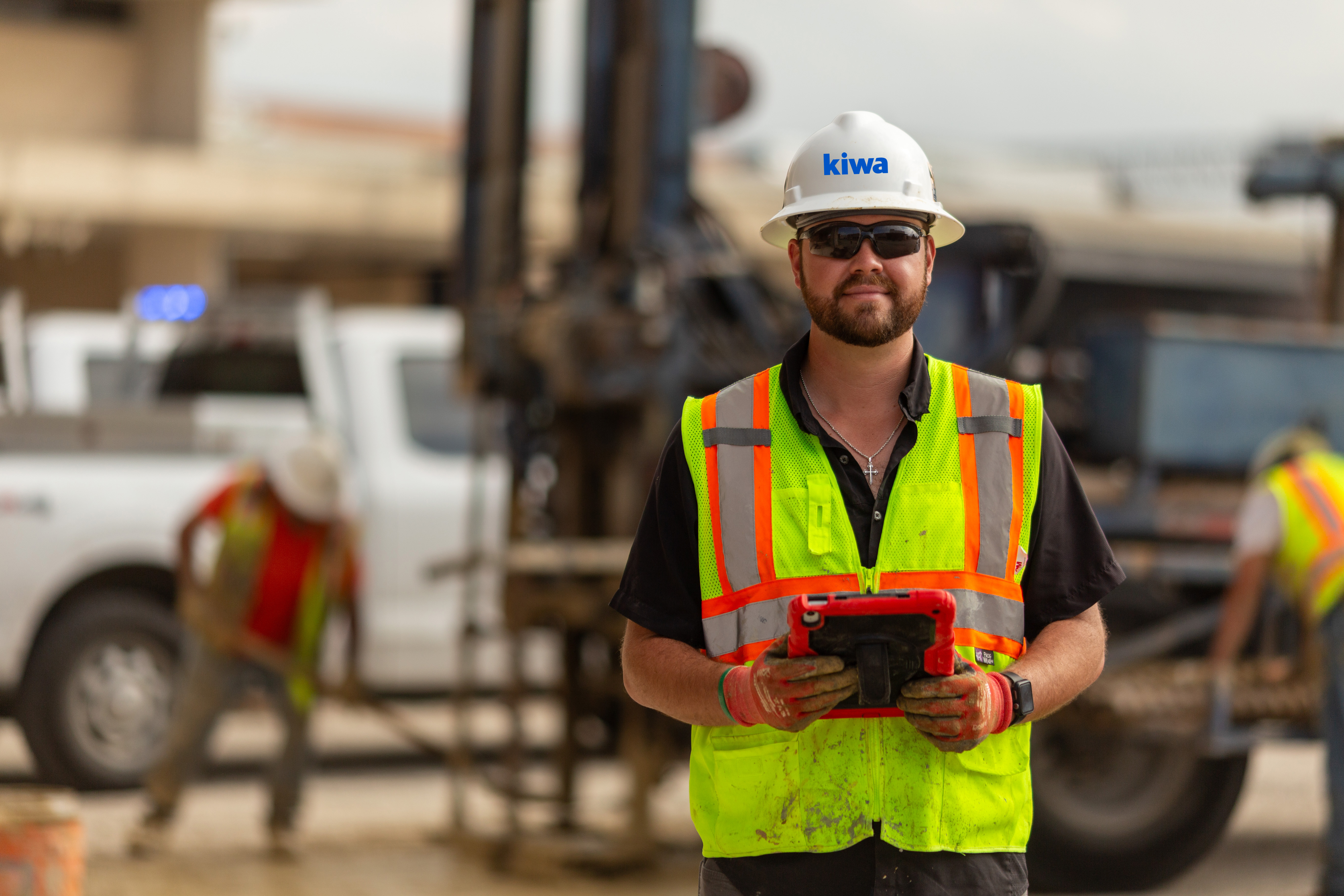 A Raba Kistner employee in a high-visibility vest and hard hat holding a tablet at a construction site, with vehicles and workers in the background.