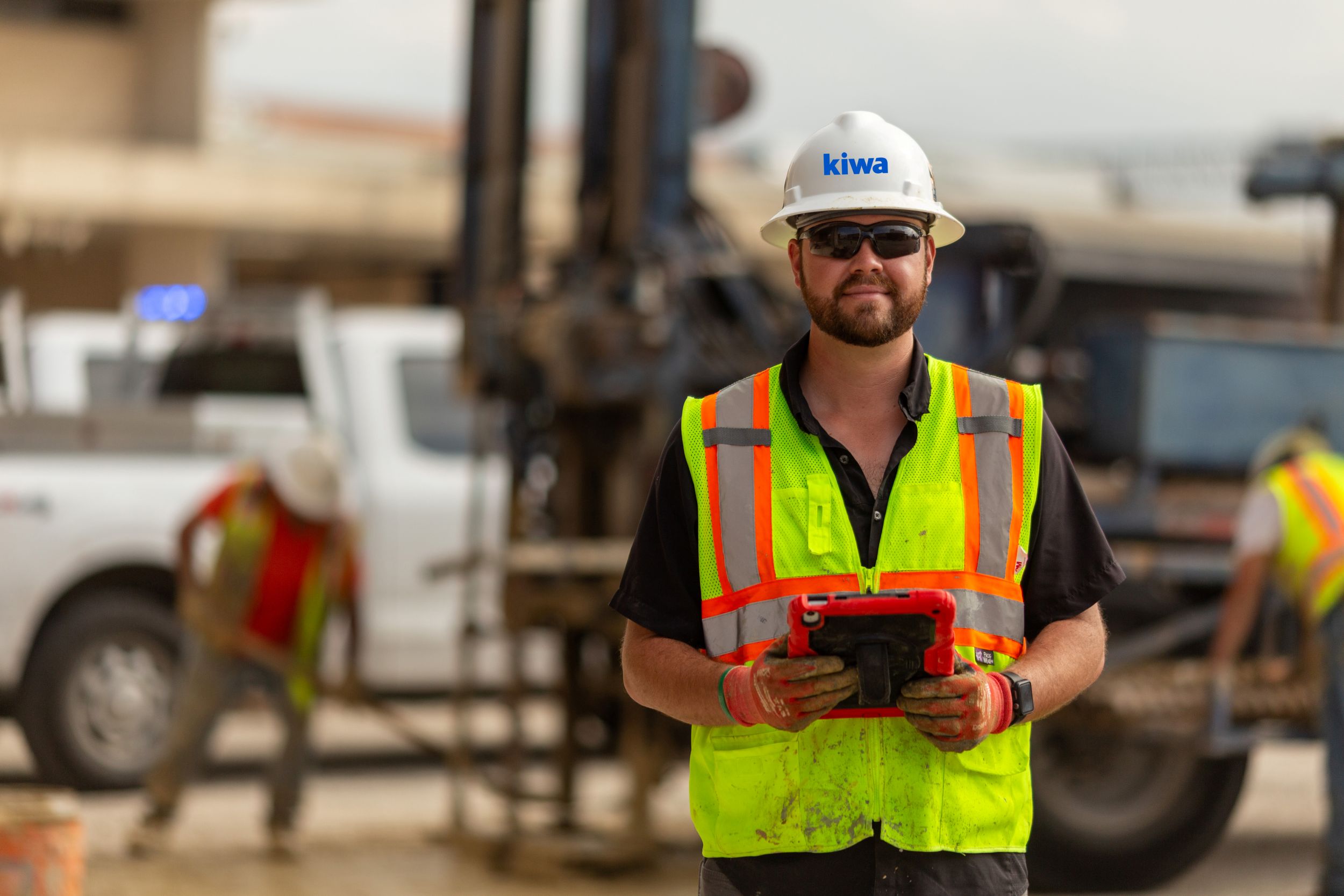 Kiwa employee in a hard hat and high-visibility safety vest holding a tablet at a job site, with heavy machinery and trucks in the background.