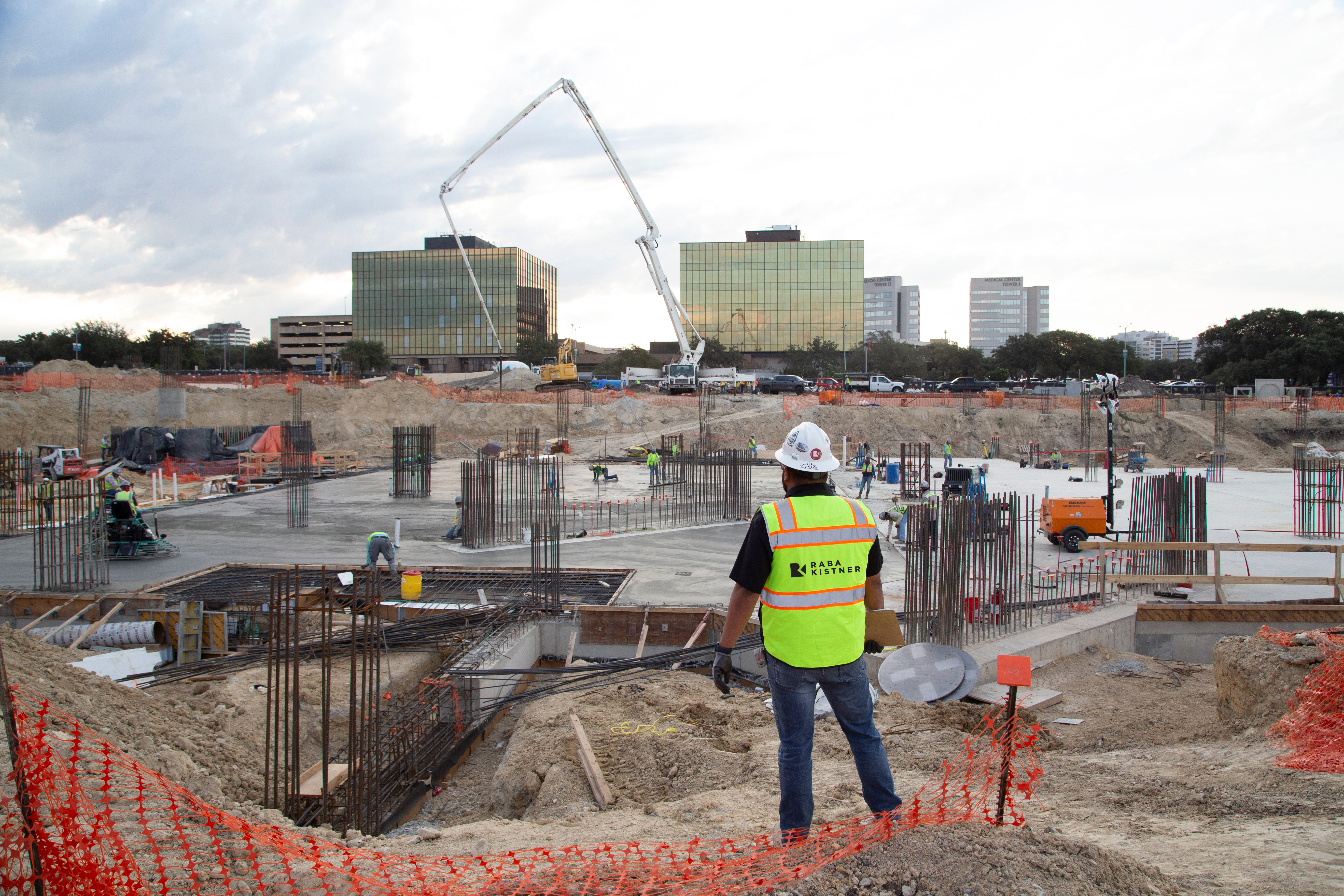 Construction site with a Kiwa Engineer in a safety vest and hard hat overlooking a large concrete foundation, rebar, and a concrete pump crane near office buildings.