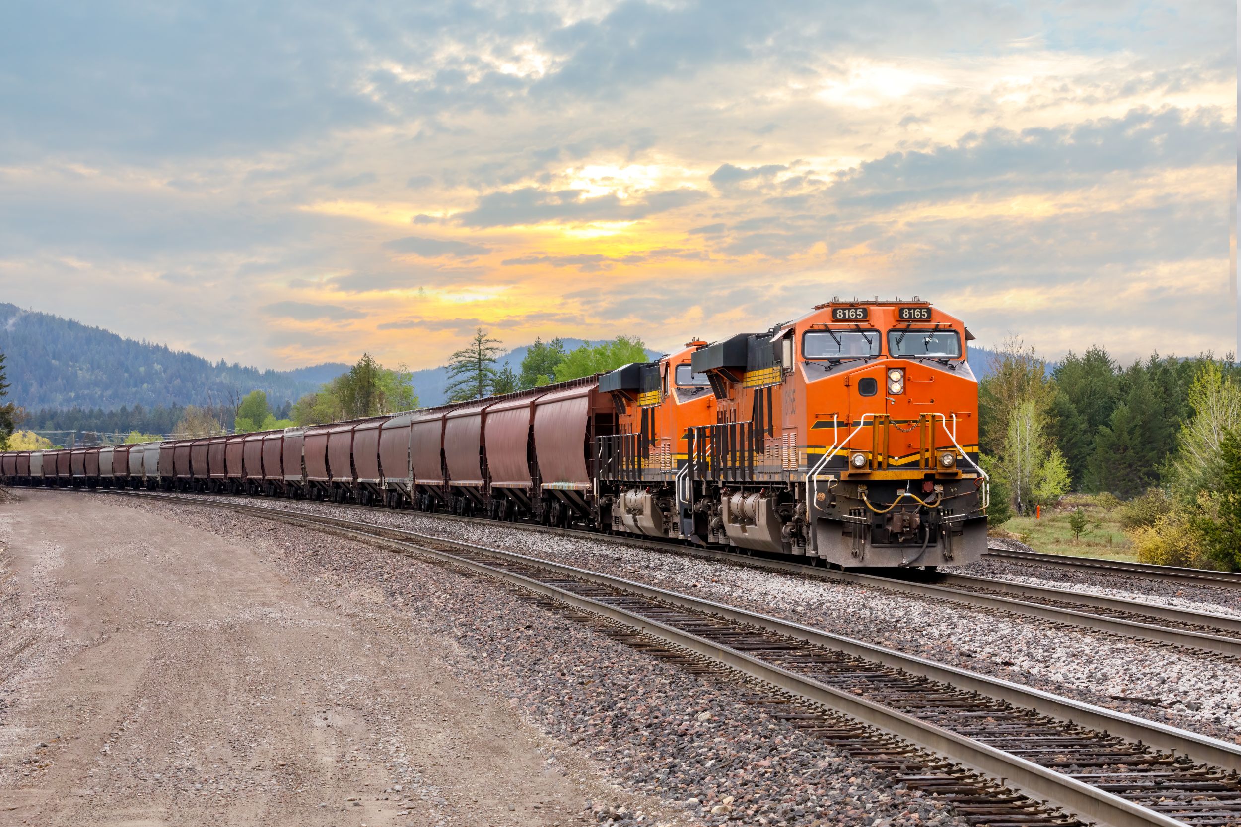 Freight train with orange locomotives pulling a long line of freight cars on railroad tracks at sunset in a rural landscape.