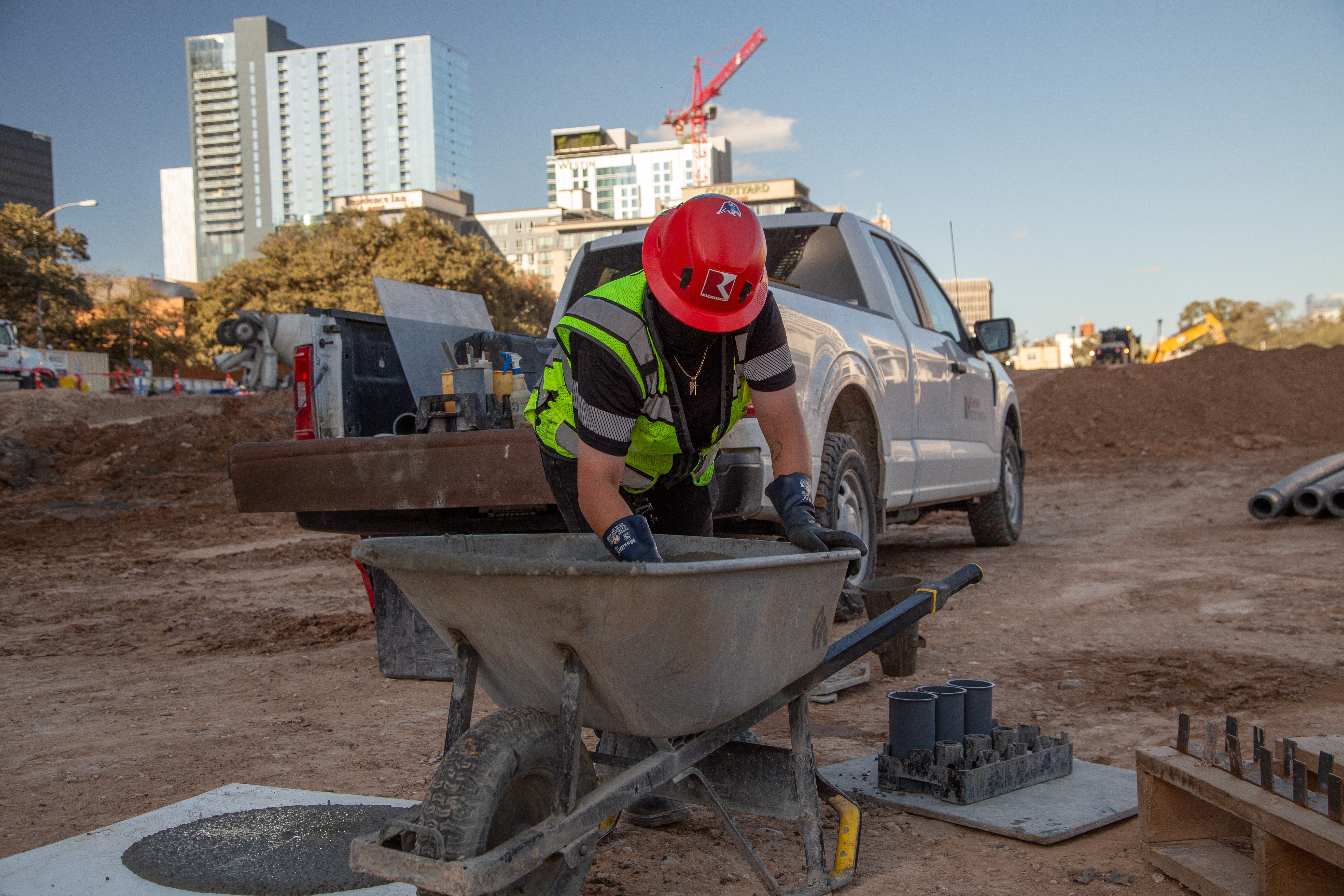 Kiwa employee in a red hard hat and safety vest mixing materials in a wheelbarrow at an urban job site beside a pickup truck.